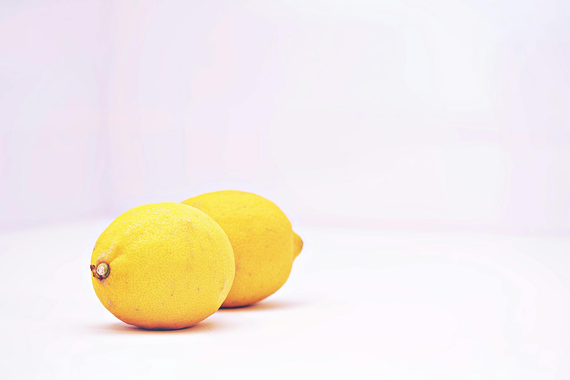 Hand mit weißen Nägeln hält eine Zitrone auf zartem rosa Hintergrund, umgeben von drei weiteren Zitronen